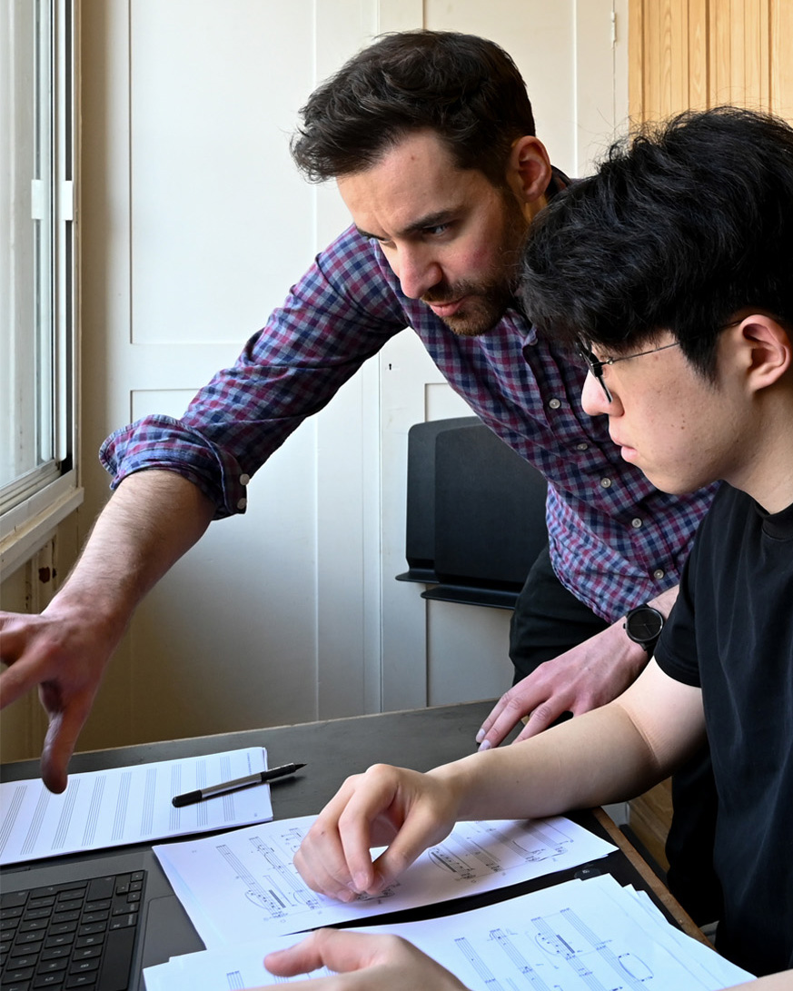 A man leaning over a younger man, directing their attention to a laptop.